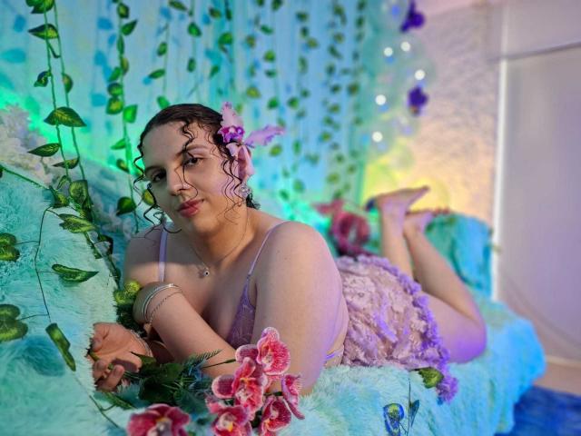 CatleyaNoire met krullend roze haar, paarse bloemen in het haar, liggend op een turquoise vacht, een roze kantjurk, roze bloemen op de voorgrond, een groene wijnrank, paars en groen licht, een blauw-witte achtergrond..