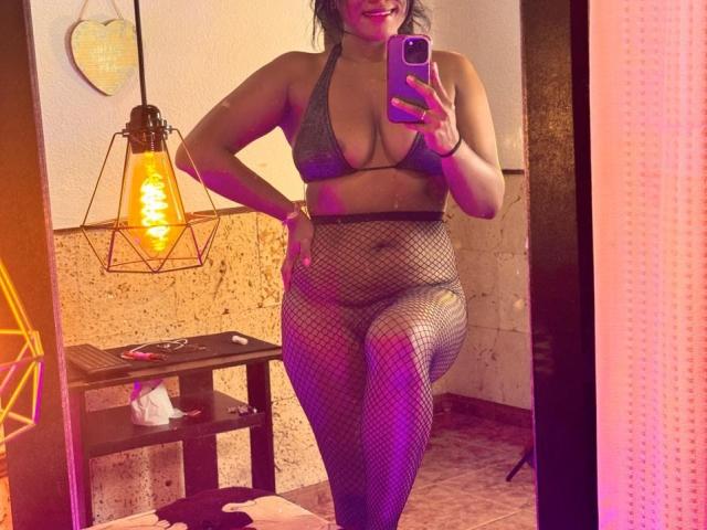 IvannaDuran in a mirror selfie wearing a gray bra, fishnet stockings, and a high-waisted bottom, showing her tits and ass. She's holding a phone, smiling, with purple lighting.