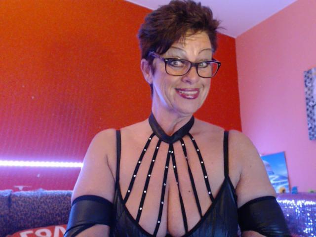 Bettina with short brown hair and glasses, wearing a black strappy top that exposes her large tits, and black arm bands, smiling at the camera.