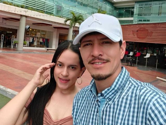 Roco has a cute chick with big tits and long black hair, wearing a pink top, standing next to a dude with a white cap and blue check shirt.