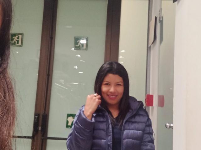 Any28-hot with medium tits, brown skin, black hair, smiling, wearing a blue puffy jacket, standing in a glass door building, hand near mouth, another person's long hair on left.