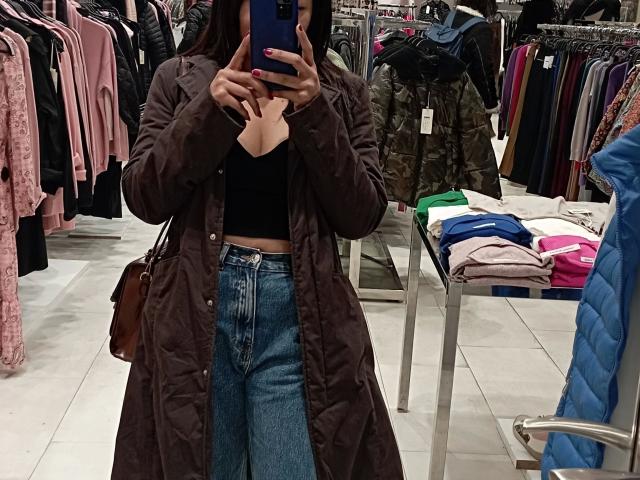 Any28-hot with brown coat, black top, and blue jeans, taking a mirror selfie in a store, showing her faceless upper body and holding a blue phone.