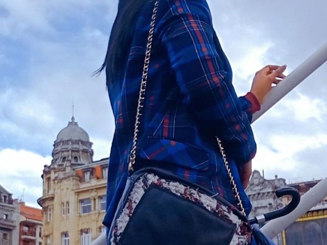 Any28-hot in a blue plaid shirt, black lace-trimmed purse, and chain strap, holding a railing with a cloudy sky and historic building in the background.