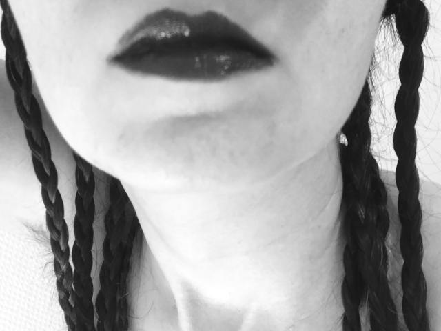 I capelli neri trecciati di AlexandraSweet, il rossetto scuro e lucido, e la pelle chiara leggermente lentigginosa, con il collo visibile e una clavicola appena accennata. Scatto in primo piano in bianco e nero..