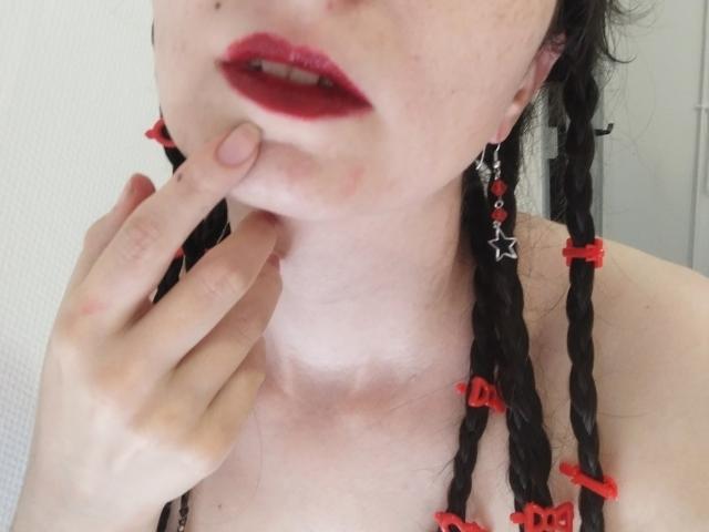 AlexandraSweet met zwarte vlechten, rode lippenstift en rode haarbandjes, die aan haar kin zit, met sterrenoorringen, bleke huid, lichte sproeten en een subtiele tatoeage op haar nek..