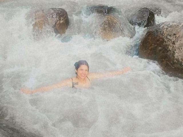 SofyaLane with wet hair, arms out, smiling in rocky river, white water swirling around her, small breasts visible, standing in current, surrounded by large brown rocks.