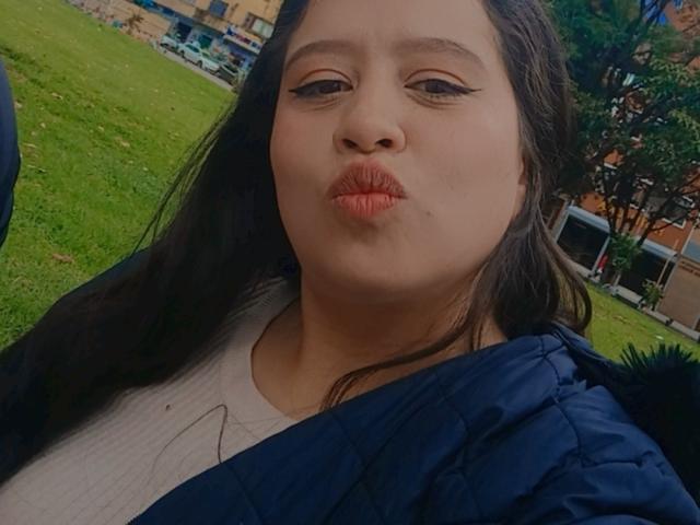 CharlloteJones with pouty lips, wearing a blue quilted jacket and white shirt, has long brown hair, brown eyes, and is making a kissing face. Her pussy is not visible.