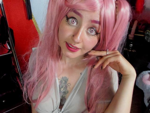 MeiJaisen with pink hair, wide purple eyes, and a nose ring, smiling at the camera, touching her cheek. Visible tattoo on her chest. She's wearing a grey top.