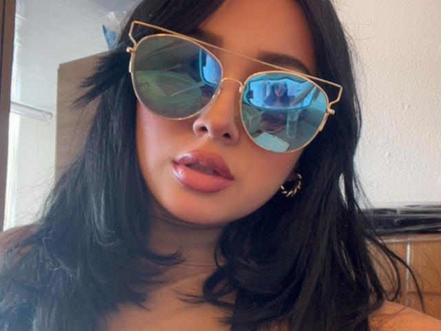 LillyTaylor with shiny pink lips, black wavy hair, and gold-rimmed reflective sunglasses, wearing gold hoop earrings, looking at camera. Reflections in glasses show blue room.