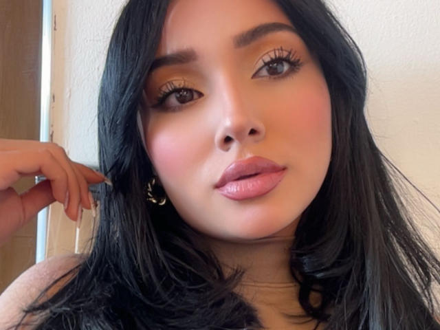 LillyTaylor has shiny black hair, full pink lips, and brown eyes with long eyelashes. She's wearing gold hoop earrings and has a hand near her face.