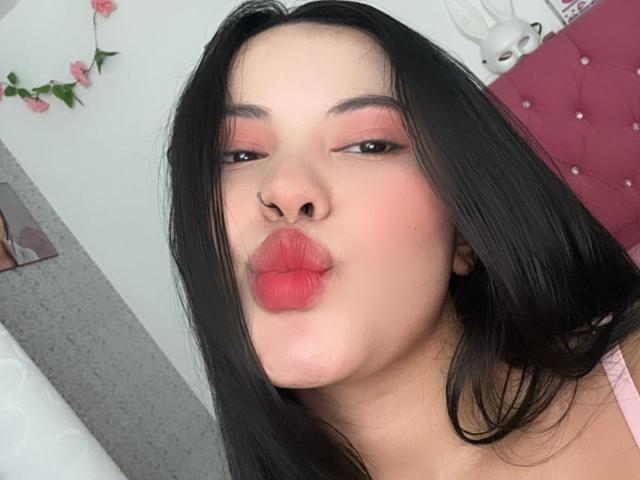 ArianaSmith69 with black hair, pink eyeshadow, and puffy red lips, puckering up. She's wearing a pink top, looking cute, with a nose ring.