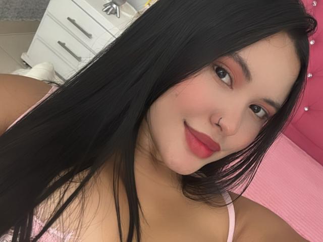 ArianaSmith69 with long black hair, pink lipstick, and a nose ring, smiling at the camera in a pink bed with a white dresser in the background. Boobs visible.