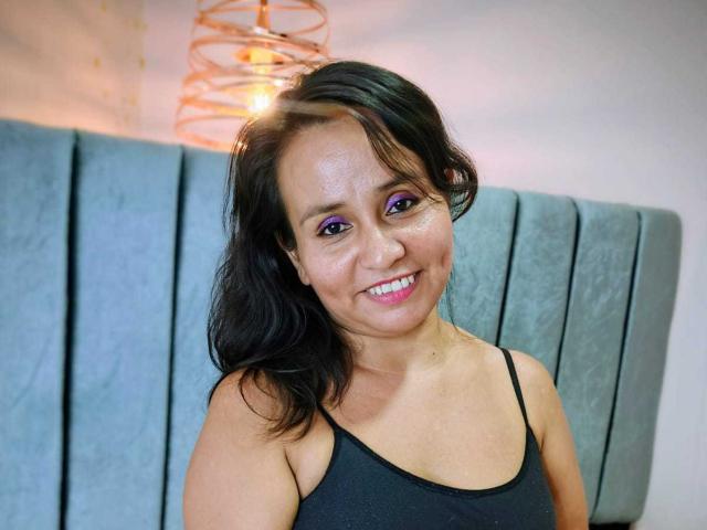 DeboraOriz heeft glanzende paarse oogmake-up, zwart haar en glimlacht met roze lippenstift, ze draagt een zwart tanktop, waardoor haar kleine borsten zichtbaar zijn, en zit tegen een grijze, beklede hoofdbord aan..