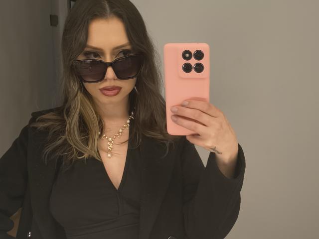 ElizaCanterville with brown wavy hair, wearing black sunglasses, black blouse, black blazer, gold necklace, and pink iPhone, taking a mirror selfie, showing her pouty red lips.