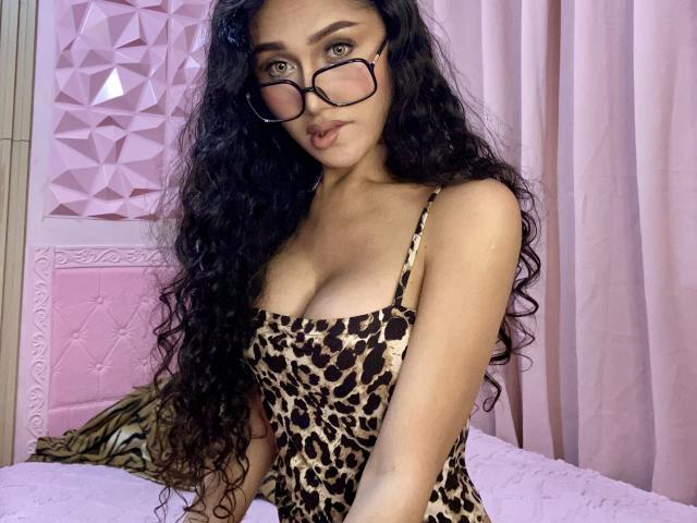 TheaLux with long curly black hair, wearing glasses and a leopard print dress, showing her boobs, standing by a pink bed with a zebra pillow.