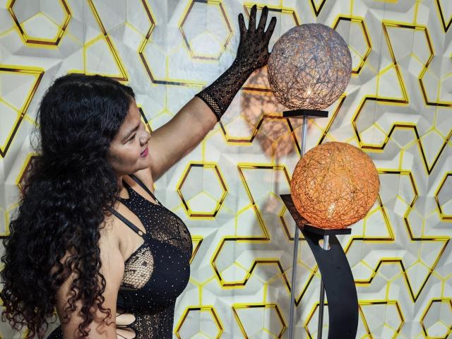 KattaleyasSex with long curly black hair, wearing a black lace dress and matching gloves, touches two ornate hanging lamps with her right hand, showing her tits.