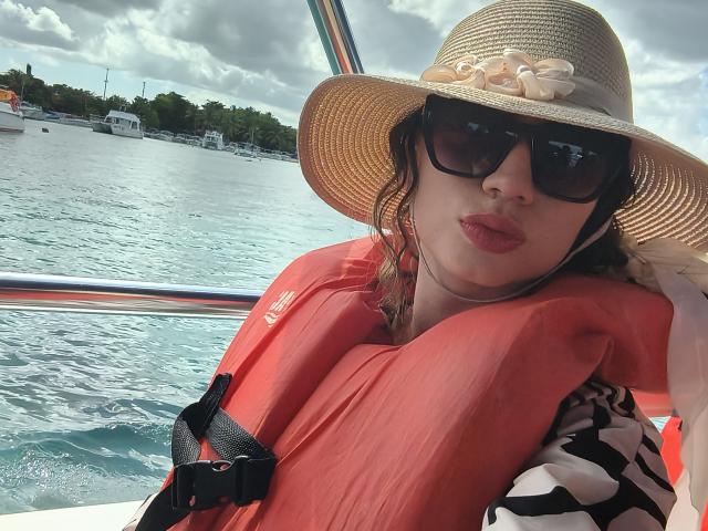 ElizaCanterville with wavy brown hair, wearing a straw hat, black sunglasses, red life jacket, and black-and-white striped shirt, on a boat, showing her pouty lips.