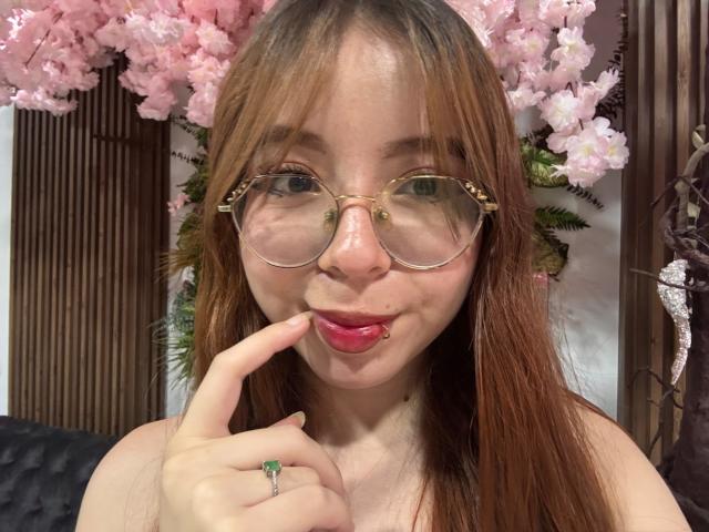 KattyHards con labbra rosa gonfie, rossetto rosso e occhiali, che morde il dito con un sorriso carino, indossando un anello verde, con fiori di ciliegio rosa sullo sfondo..