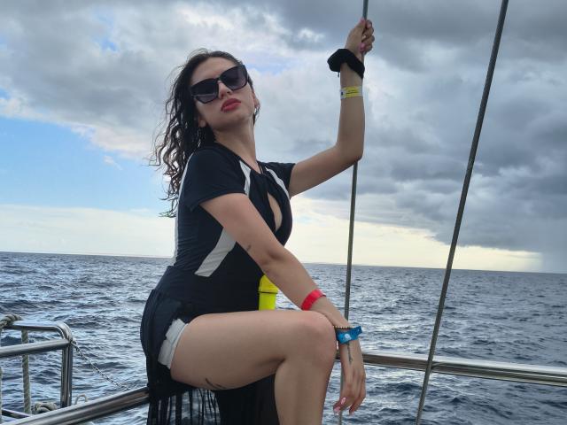 ElizaCanterville with curly dark hair, wearing black dress, sunglasses, and wristbands, gripping sailboat rope, showing her ass, on a cloudy ocean. Her red lipstick contrasts the grey sky.
