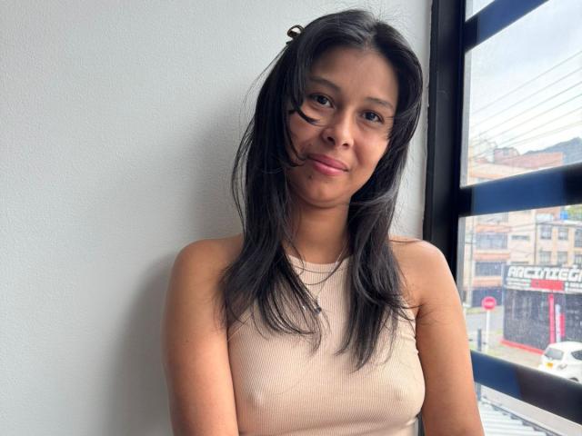 VanessaHayle with straight black hair, small tits, and brown skin, wearing a beige sleeveless top, standing near a window, smiling slightly. Her nipples are slightly visible.