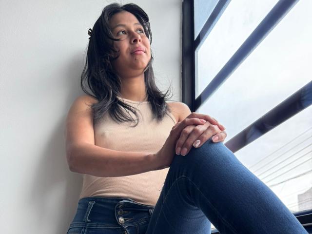 VanessaHayle with black hair, tan top, and blue jeans, sitting by a window, looking up, hands on knee, slight smile, natural light, casual, relaxed, medium shot.