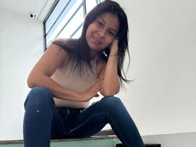 VanessaHayle is sitting with her knees up, wearing a white tank top and blue jeans, smiling and resting her head on her hand. Her ass is slightly visible.