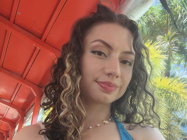 ElizaCanterville has curly brown hair with blonde highlights, black eyeliner, pink lipstick, and a pearl necklace, standing under a red roof with palm trees in the background.