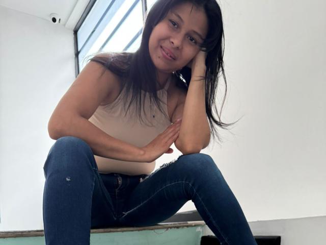 VanessaHayle with long dark hair, smiling, wearing a white tank top and blue jeans, sitting with legs apart, hand on cheek, in a bright room with a window.