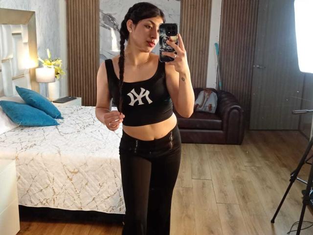 TatianaMorelli with black braided hair, wearing a black crop top with white NY logo, black pants, standing in a bedroom, taking a mirror selfie with a phone.