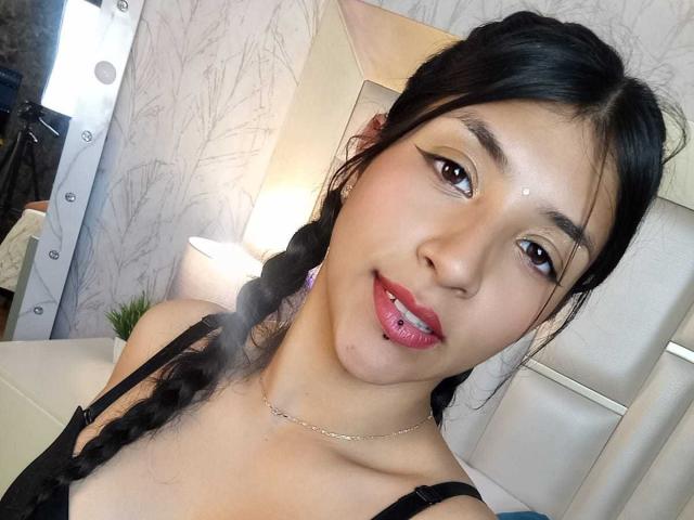 TatianaMorelli has dark hair in braids, red lipstick, and a small dot on her forehead. She's wearing a black top and a silver necklace, looking at the camera.