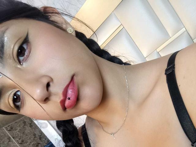 TatianaMorelli with glossy brown eyes, black braided hair, shiny gold eyeshadow, pierced tongue, pink lipstick, silver necklace, black strap, light skin, small earrings, white wooden chair.