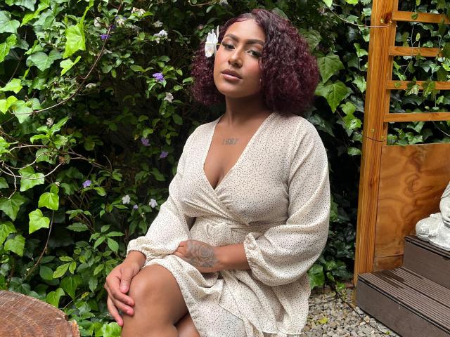 AmberQueeny, avec ses cheveux violets bouclés, sa peau bronzée et ses tatouages, portant une robe beige texturée, assise les mains sur ses genoux, montrant sa poitrine et affichant un sourire discret..