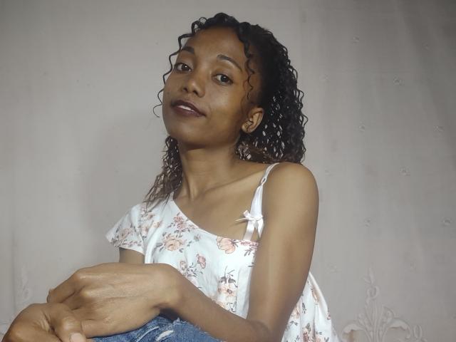 VictorineStone with curly brown hair, wearing a floral white top and denim jeans, smiling at the camera, hands resting on her lap, showing her small tits and dark brown skin.