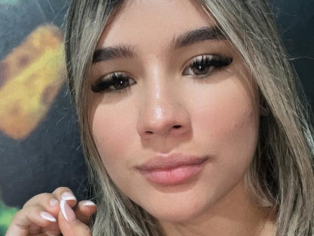 SashaPolet has light brown skin, long grayish-blonde hair, and is looking at the camera with full pink lips and thick, long eyelashes. Her hand is near her face.