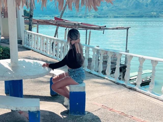 SashaPolet with long brown hair, black top, denim shorts, squatting on blue and white bench, hands on white railing, looking at calm blue water, under thatched roof.
