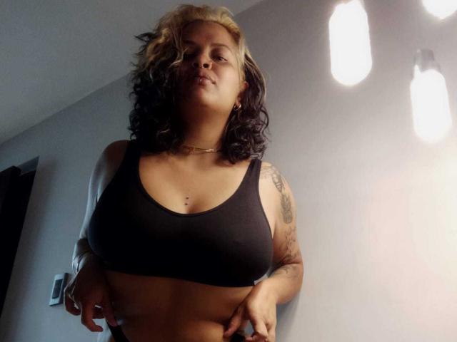 MariGonz has dark and blonde curly hair, a black sports bra showing her tits, tattoos on her arm, and a gold necklace, standing in a dimly lit room.