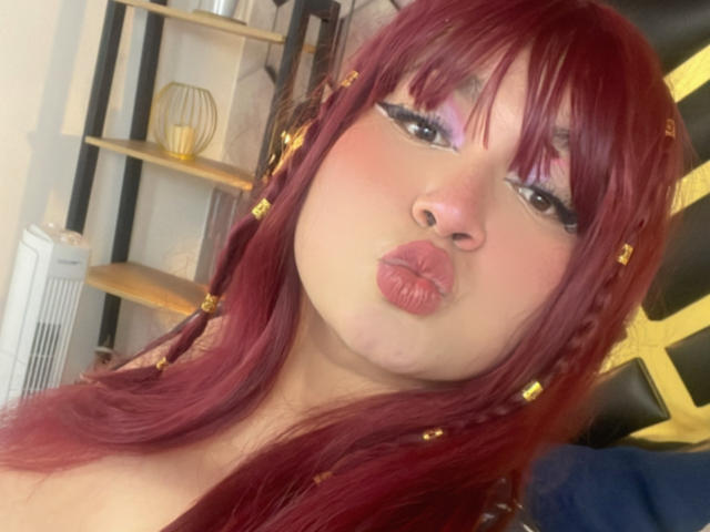 LillyMayer with red hair, puffy pink lipstick, and gold hair clips, making a pouty face, showing off her pink cheeks and subtle nose. Her tits are partially visible.