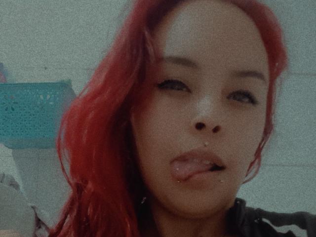 AlanaRoth has red hair, slightly parted lips with a small piercing on her upper lip, and is sticking out her tongue. Her eyes are half-open.
