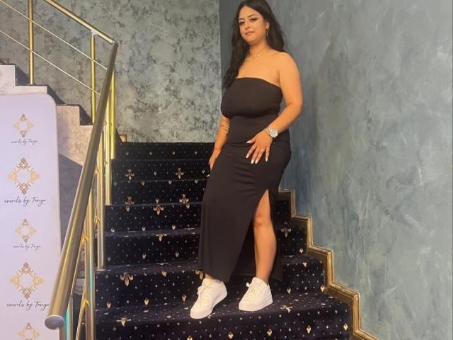 GloriaHaylee stands on a staircase, wearing a black strapless dress with a high slit, white sneakers, and a watch, with her hands on her ass.