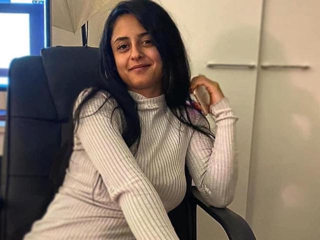 GloriaHaylee, sitting in a black chair, wearing a ribbed white turtleneck, has long black hair and is smiling slightly with one hand touching her hair.