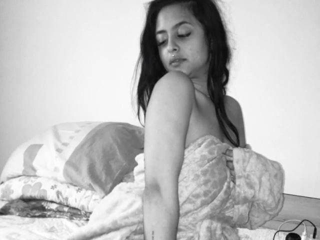 GloriaHaylee with long dark hair, eyes closed, sitting on bed, covered in a floral-patterned blanket, showing her bare shoulders and a small tattoo on her right arm.