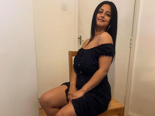 GloriaHaylee, sitting on a wooden chair, wearing an off-shoulder black dress, showing her ass, with long black hair, and a subtle smile, in a simple room.