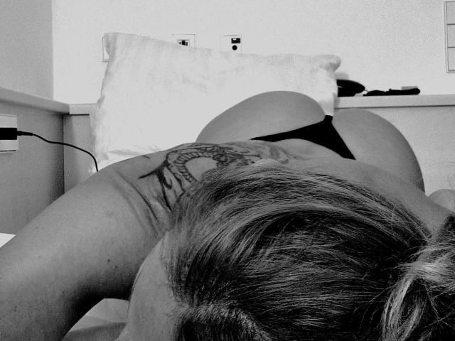 Irina-Faust-hot is on all fours, showing her tattooed back and black panties, with her ass raised and blonde hair down. Black and white, top-down, slightly blurred.