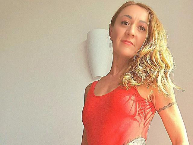 Irina-Faust, une femme séduisante aux cheveux blonds ondulés, portant un débardeur rouge, un tatouage sur le bras droit et un sourire discret, debout devant un mur uni, avec un maquillage très léger..