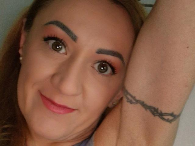 Irina-Faust-hot with a tattoo on her raised arm, brown eyes, pink lipstick, and a slight smile. She has arched eyebrows, long eyelashes, and a light skin tone.