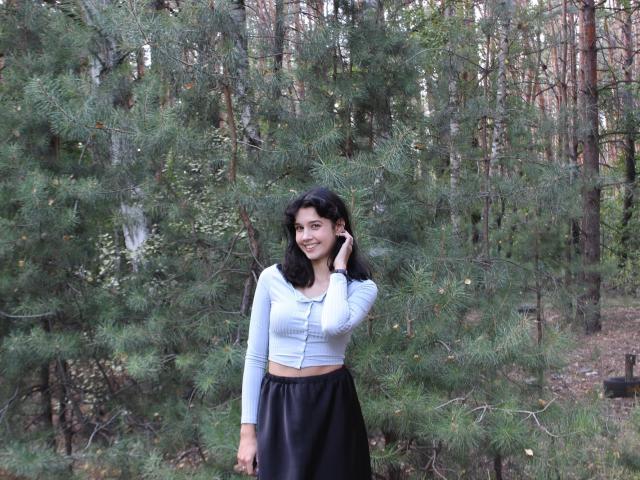 LettyCurly with black hair, wearing a light gray crop top and black skirt, standing in a forest, holding a phone to her ear, smiling.