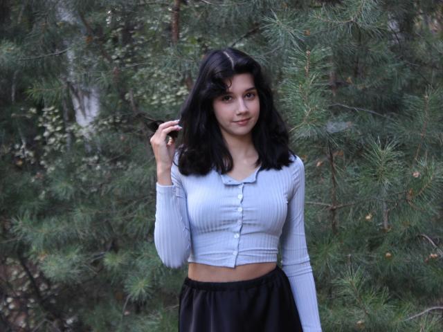LettyCurly with medium tits, wearing a light blue ribbed crop top and black skirt, standing in front of pine trees, touching her wavy black hair with a slight smile.