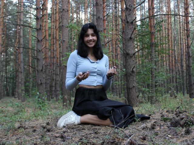 LettyCurly, with black curly hair, sits on forest floor, wearing a light blue crop top, black skirt, and white sneakers, holding small objects, smiling, showing off her tits.