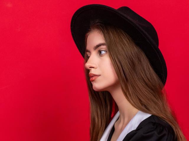 EricaDiaz with long brown hair, black hat, and pouty lips, looking to the side, wearing a black and white top, against a bright red background.