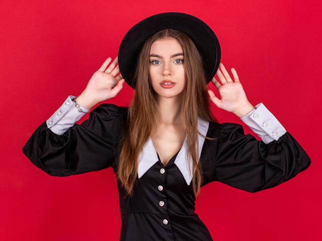 EricaDiaz with long brown hair, blue eyes, wearing a black dress with white collar, black hat, and holding it up, red background, showing her tits and ass.